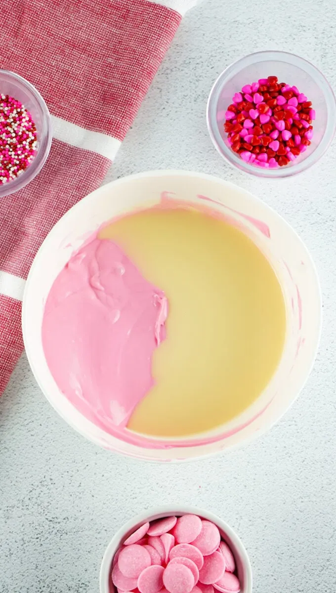 A bowl with pink and cream-colored melted candy, surrounded by small bowls of heart and sprinkle decorations, and pink candy melts. A red and white cloth is partially visible.