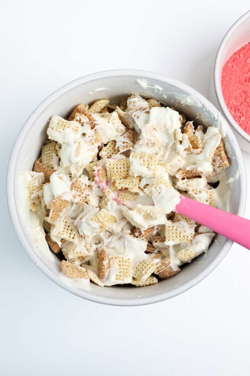 A bowl of cereal mix with white coating and a pink spatula, next to a small bowl with red powder.