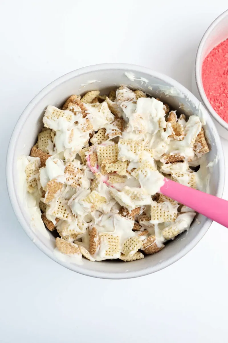 A bowl of cereal mix with white coating and a pink spatula, next to a small bowl with red powder.