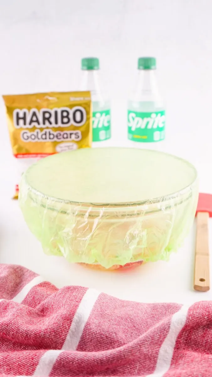 A bowl covered in green plastic sits on a table with a red and white cloth, two Sprite bottles, and a Haribo Goldbears package in the background.