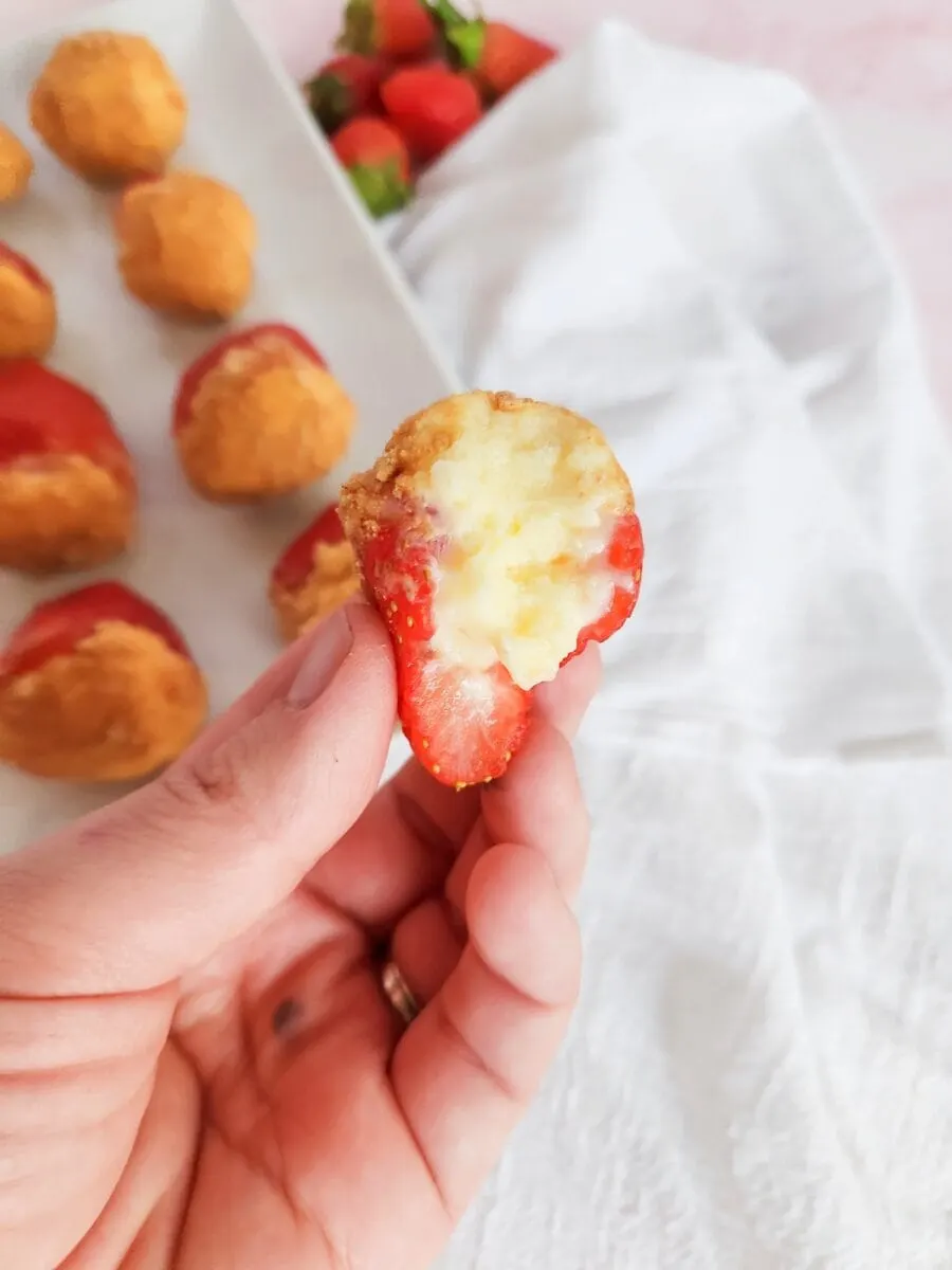 A hand holds a half-eaten strawberry filled with a creamy substance, with more strawberries on a plate in the background.