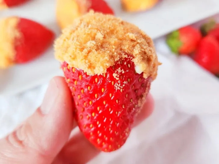 A hand holds a strawberry stuffed with cheesecake filling and topped with graham cracker crumbs against a white cloth background