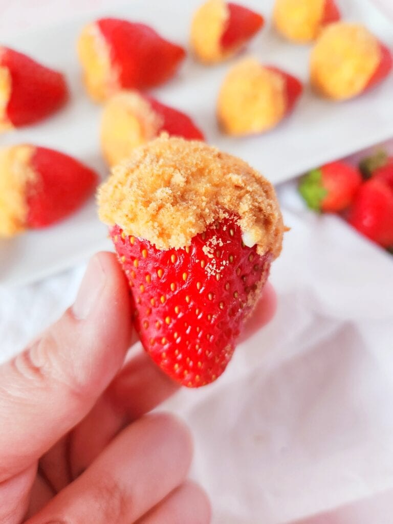 A hand holds a strawberry stuffed with cheesecake filling and topped with graham cracker crumbs against a white cloth background