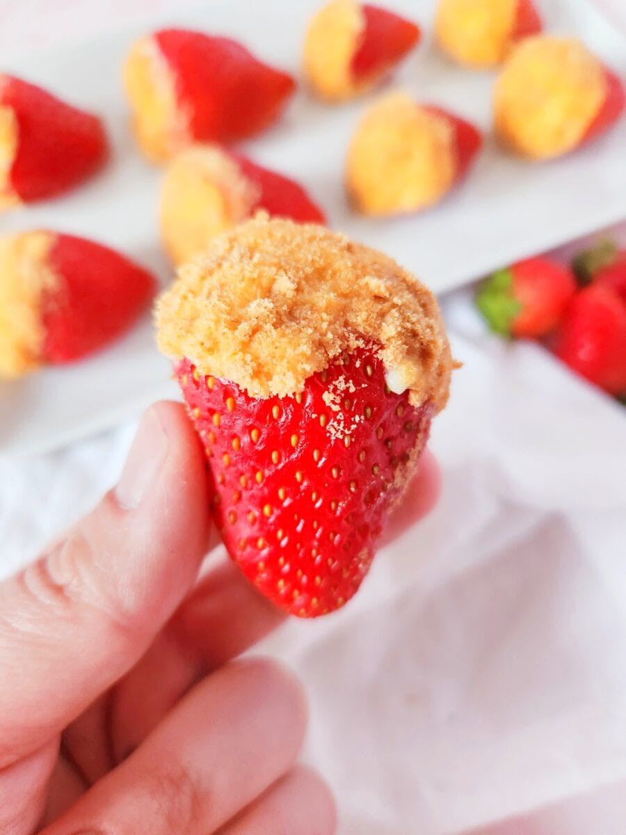 A hand holds a strawberry coated with a crumbled topping, reminiscent of cheesecake stuffed strawberries, with more similarly topped delights on a plate in the background.