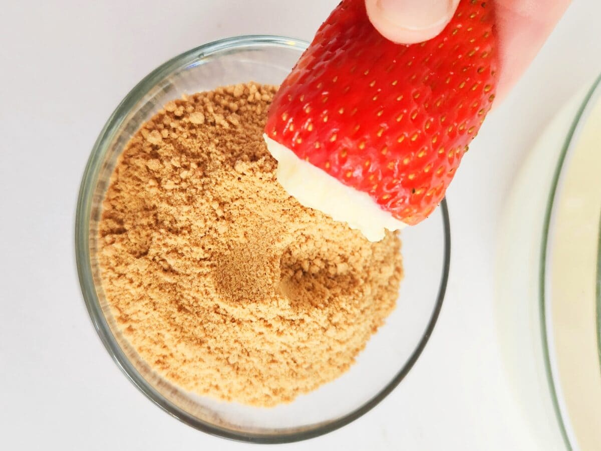 A hand holding a strawberry dipped in white chocolate above a bowl of brown sugar.