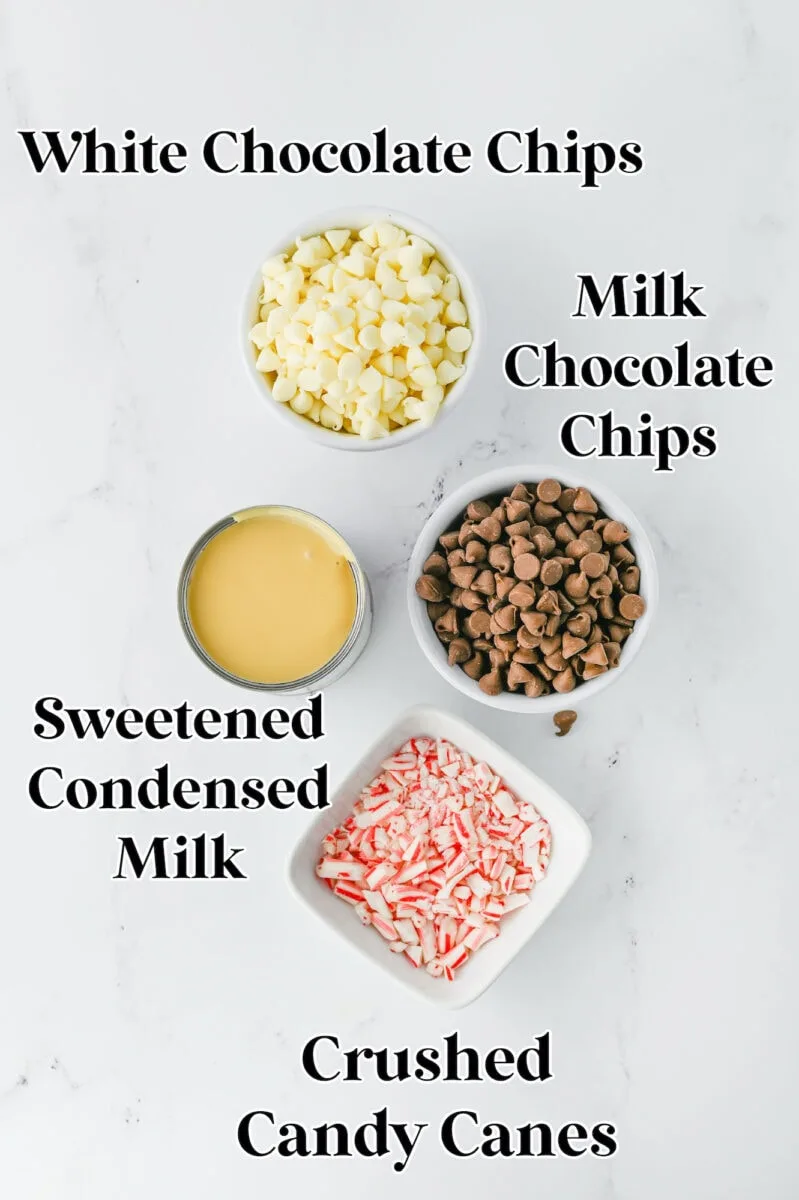 Four bowls with baking ingredients: white chocolate chips, milk chocolate chips, sweetened condensed milk, and crushed candy canes on a white surface.