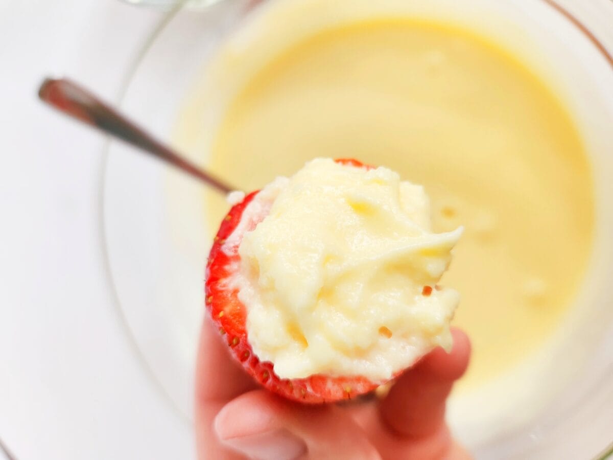 A strawberry filled with a creamy white mixture, held up by a hand over a bowl of more cream with a spoon.