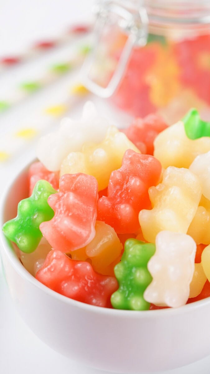 A bowl of colorful gummy bears in green, red, yellow, and white, with a glass jar and more gummies in the background.