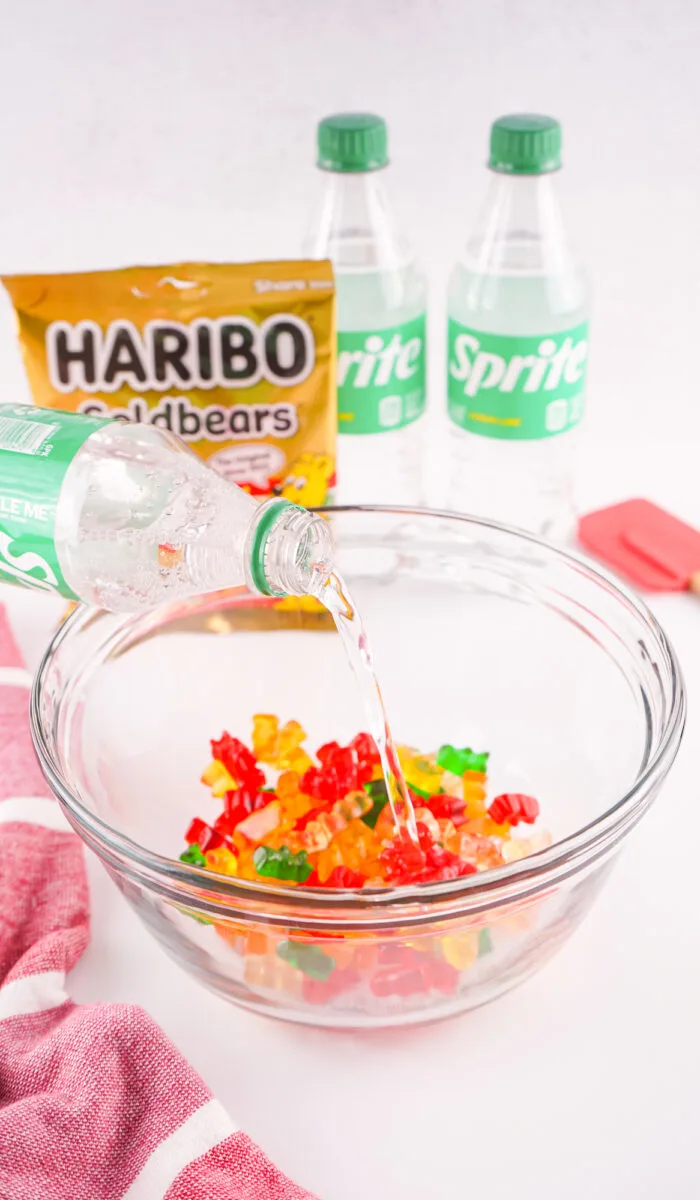 Sprite being poured over gummy bears in a glass bowl, with more Sprite bottles and a Haribo Goldbears pack in the background.