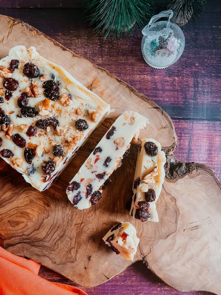 Sliced nut and berry fudge from homemade fudge recipes sits on a wooden board, with a decorative bauble and evergreen sprigs in the background.