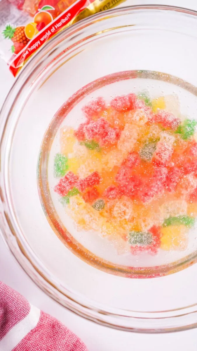 A glass bowl filled with gummy bears dissolving in a clear liquid. A packet of gummy bears is partially visible in the background.