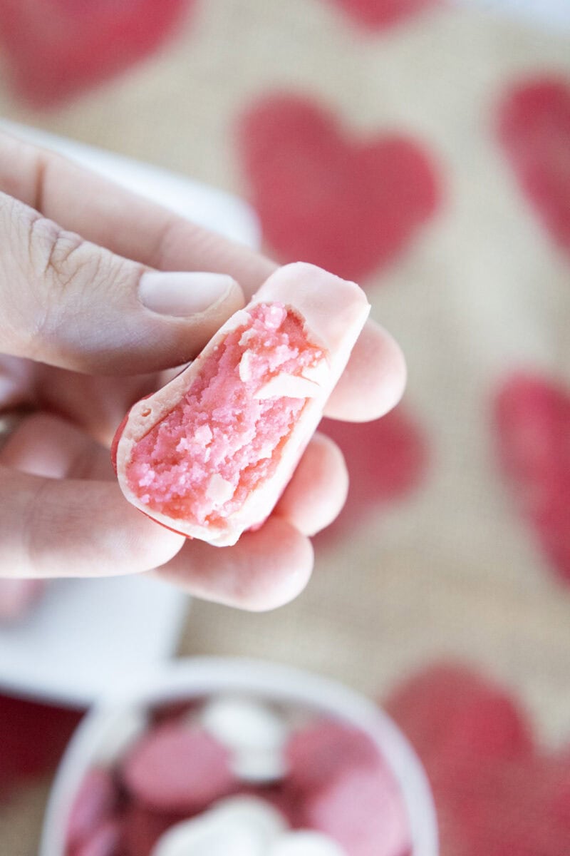 A hand holds a bitten pink candy with a textured interior. Red heart shapes are visible in the blurred background.