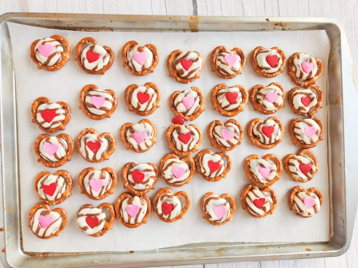 A baking tray with pretzel treats topped with chocolate, and heart-shaped decorations in red, pink, and white.