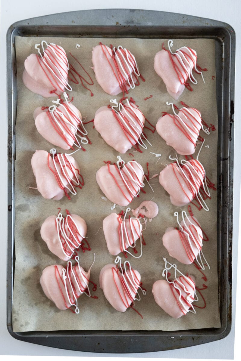 Heart-shaped cookies with pink icing and red and white drizzle on a parchment-lined baking sheet.