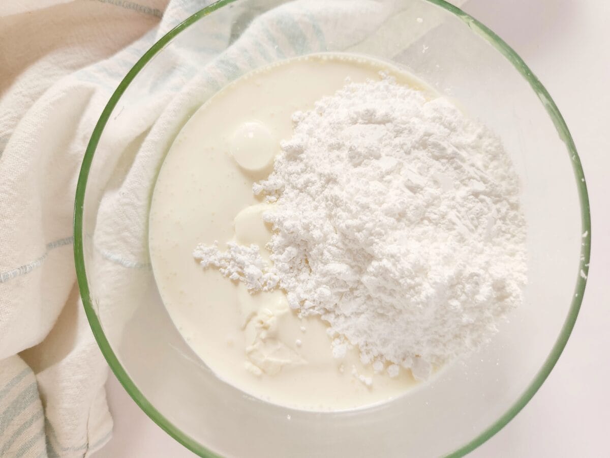 A glass bowl containing milk and a mound of flour on top, with a striped cloth to the side.