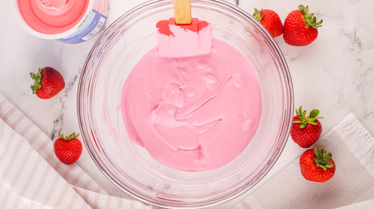 A glass bowl with pink yogurt and a red spatula, surrounded by whole strawberries on a marble surface.