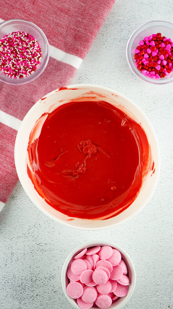 A bowl of red melted chocolate is surrounded by small bowls of colorful sprinkles and pink chocolate discs on a white surface with a red and white towel.