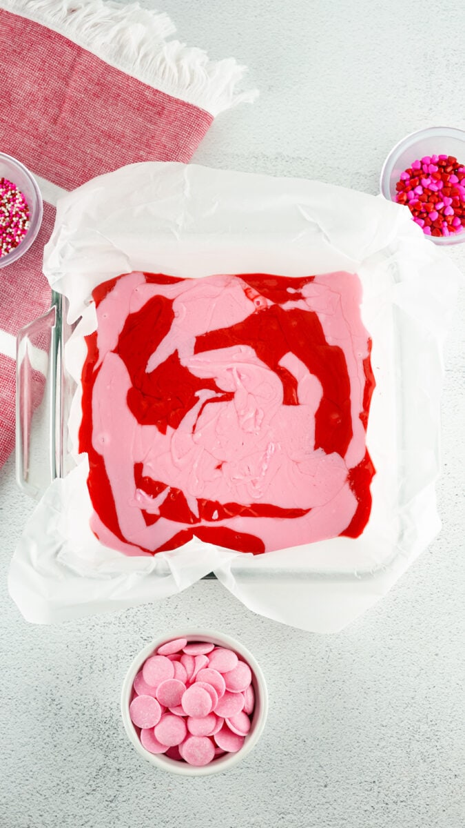 A baking pan with pink and red swirled batter sits on a countertop, surrounded by bowls of pink and red sprinkles and candy melts, with a red and white cloth nearby.