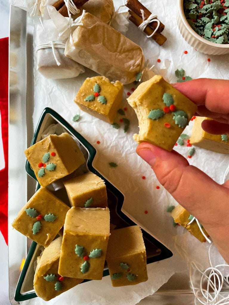 A person holds a piece of fudge adorned with holly leaf decorations, surrounded by an array of homemade treats, showcasing the perfect outcome of cherished fudge recipes.