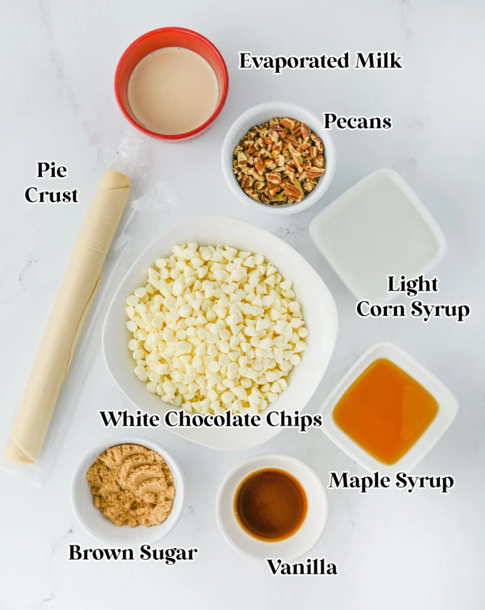 Ingredients for a pecan pie fudge delight rest on a white surface: pie crust, evaporated milk, pecans, light corn syrup, white chocolate chips, maple syrup, brown sugar, and vanilla.