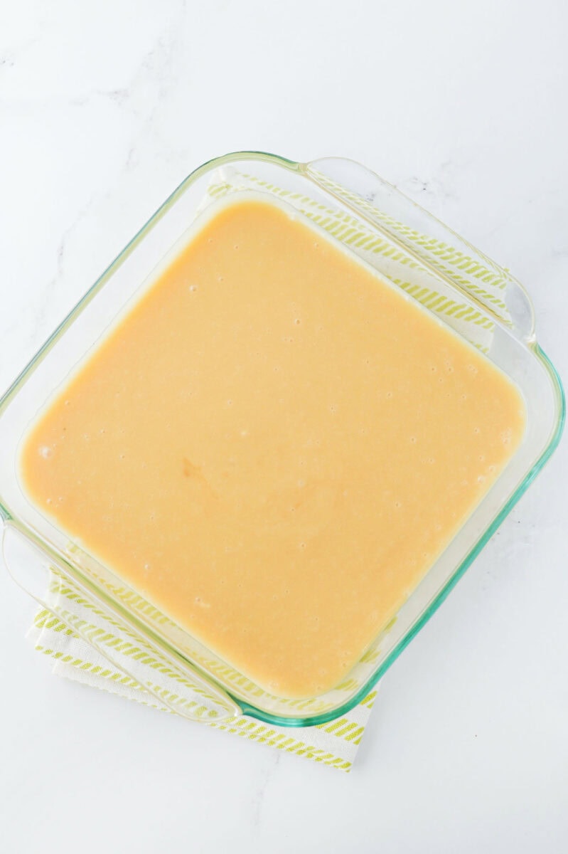 A glass container brimming with smooth, light beige batter is set atop a striped cloth, viewed from above—a perfect prelude to pecan pie fudge indulgence.