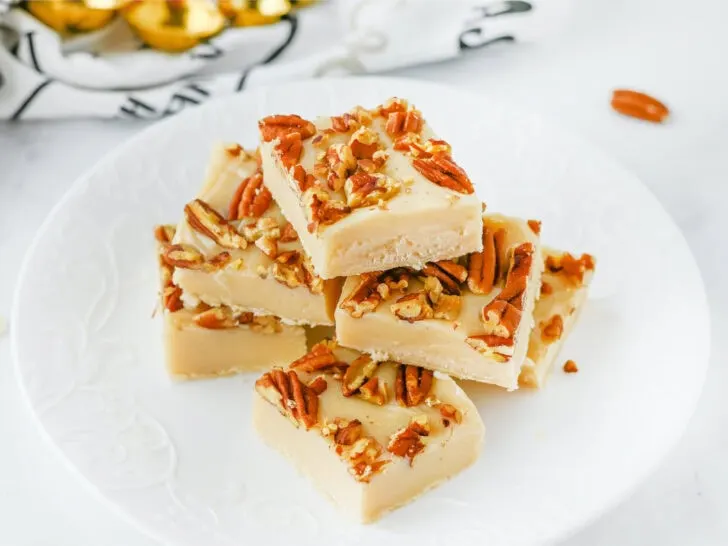 Four pieces of pecan pie fudge stacked on a white plate, each adorned with crunchy nuts.