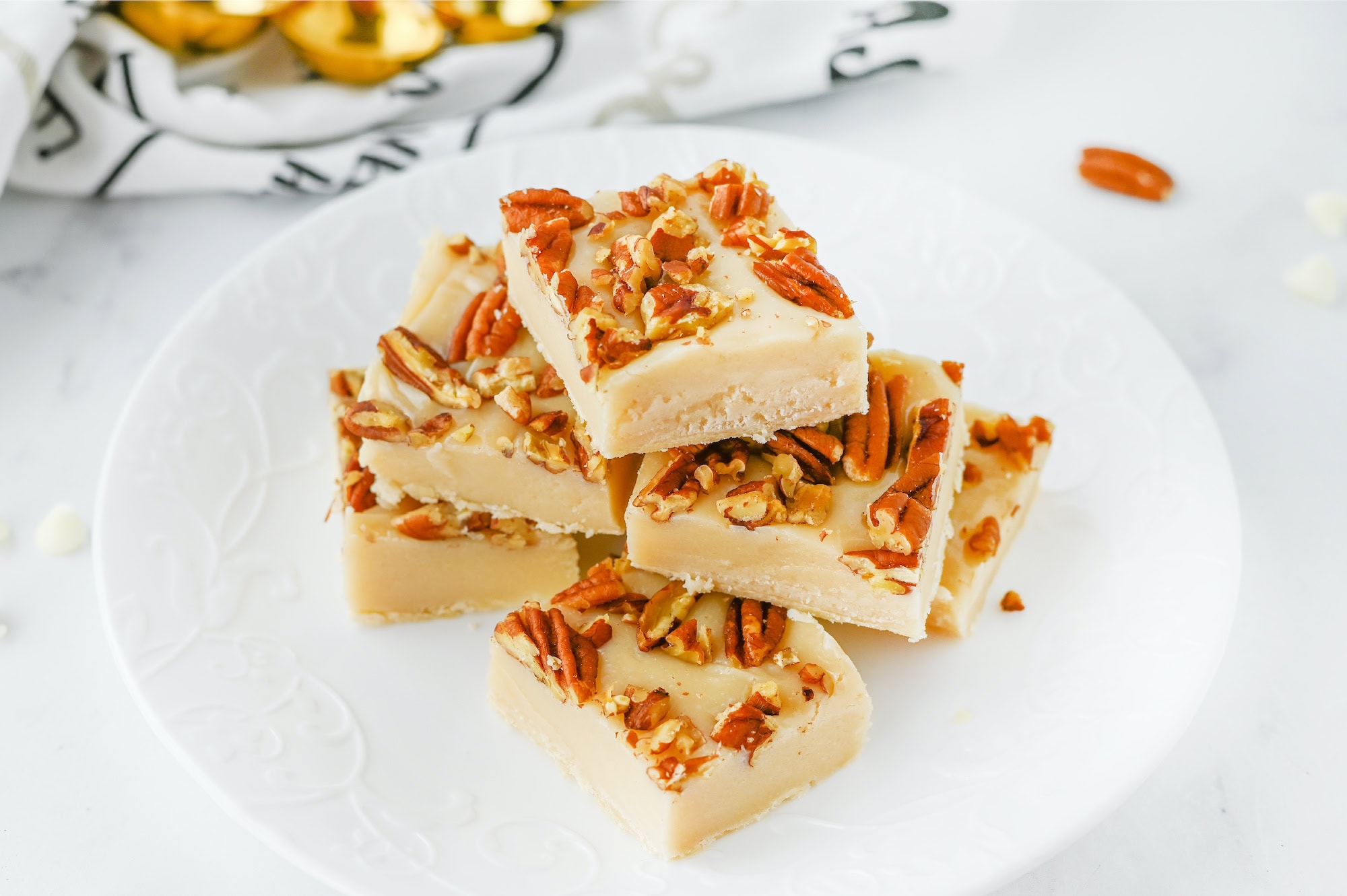 Four pieces of pecan pie fudge stacked on a white plate, each adorned with crunchy nuts.