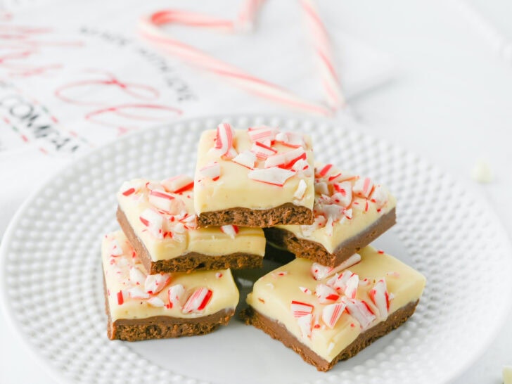 A stack of peppermint bark squares on a white plate, featuring a layer of chocolate topped with white chocolate and crushed candy canes.