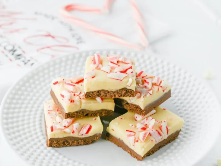 A stack of peppermint bark squares on a white plate, featuring a layer of chocolate topped with white chocolate and crushed candy canes.