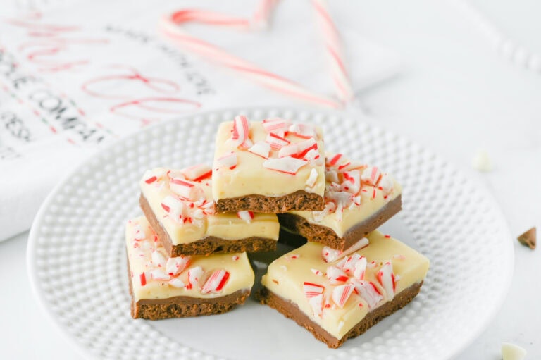 A stack of peppermint bark squares on a white plate, featuring a layer of chocolate topped with white chocolate and crushed candy canes.