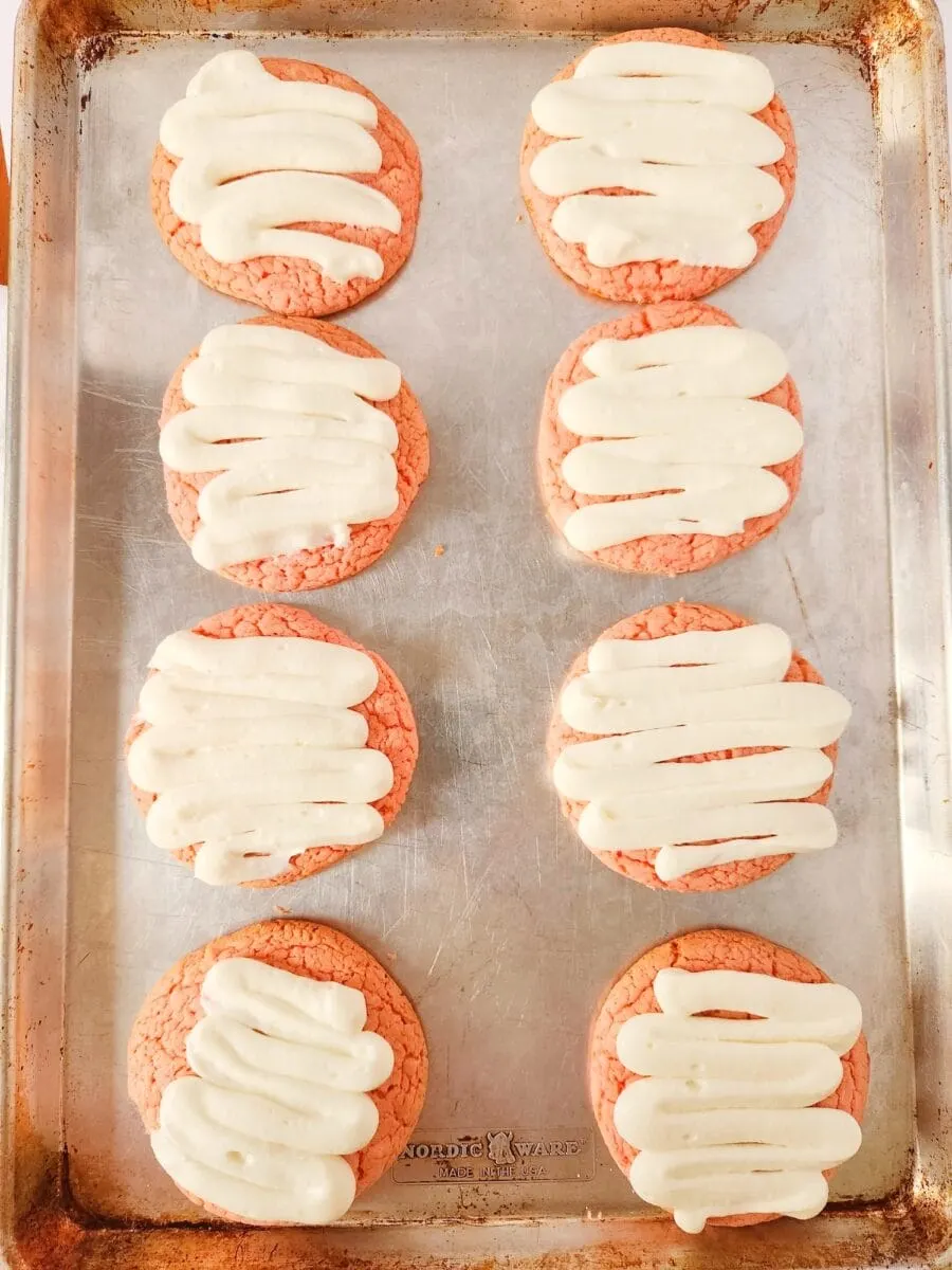 Eight pink cookies with white frosting on a baking sheet.