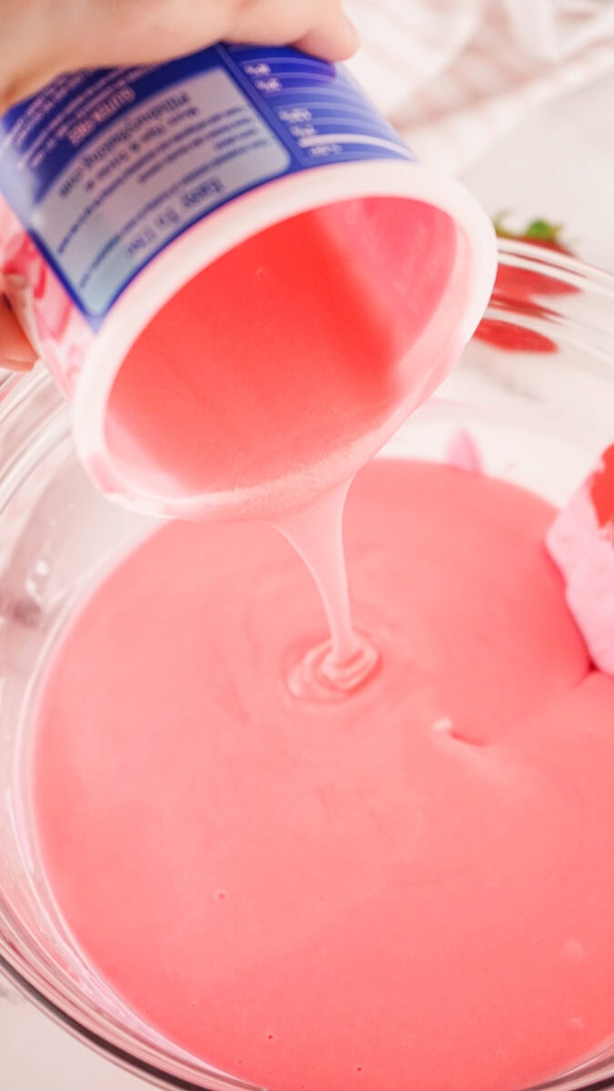 Pink batter is being poured from a container into a glass bowl.