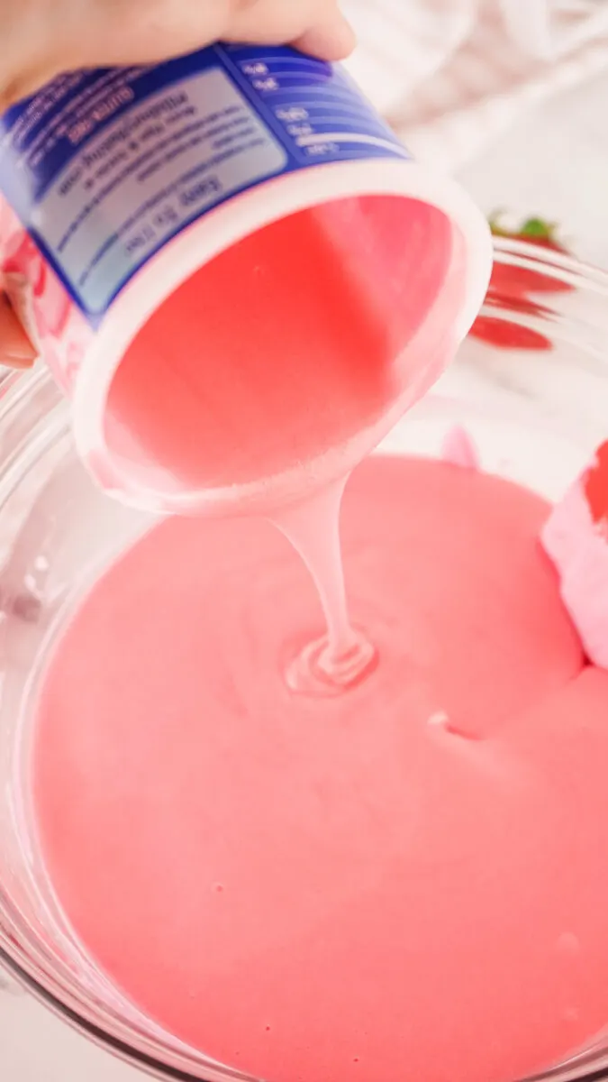 Pink batter is being poured from a container into a glass bowl.