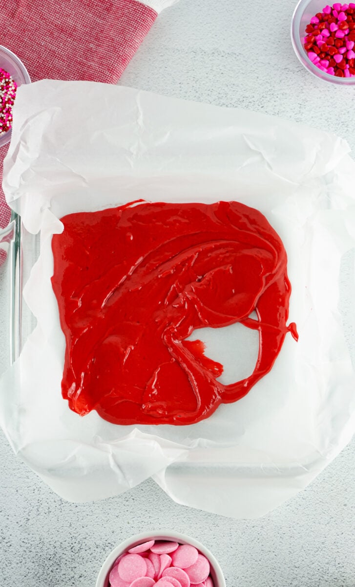 Red cake batter spread in a parchment-lined square baking dish, surrounded by bowls of pink and red sprinkles on a light gray surface.