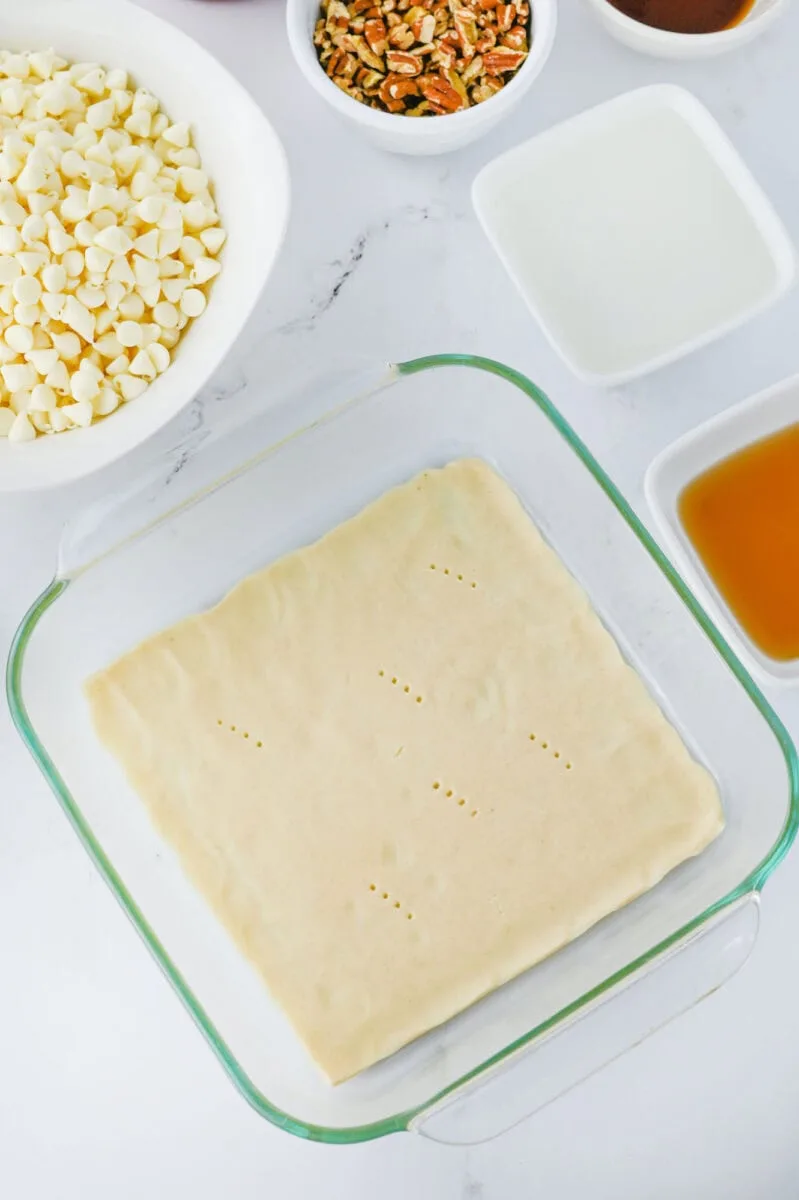 A glass baking dish with unbaked pecan pie fudge dough is surrounded by bowls of white chocolate chips, pecans, and various liquids on a pristine white surface.
