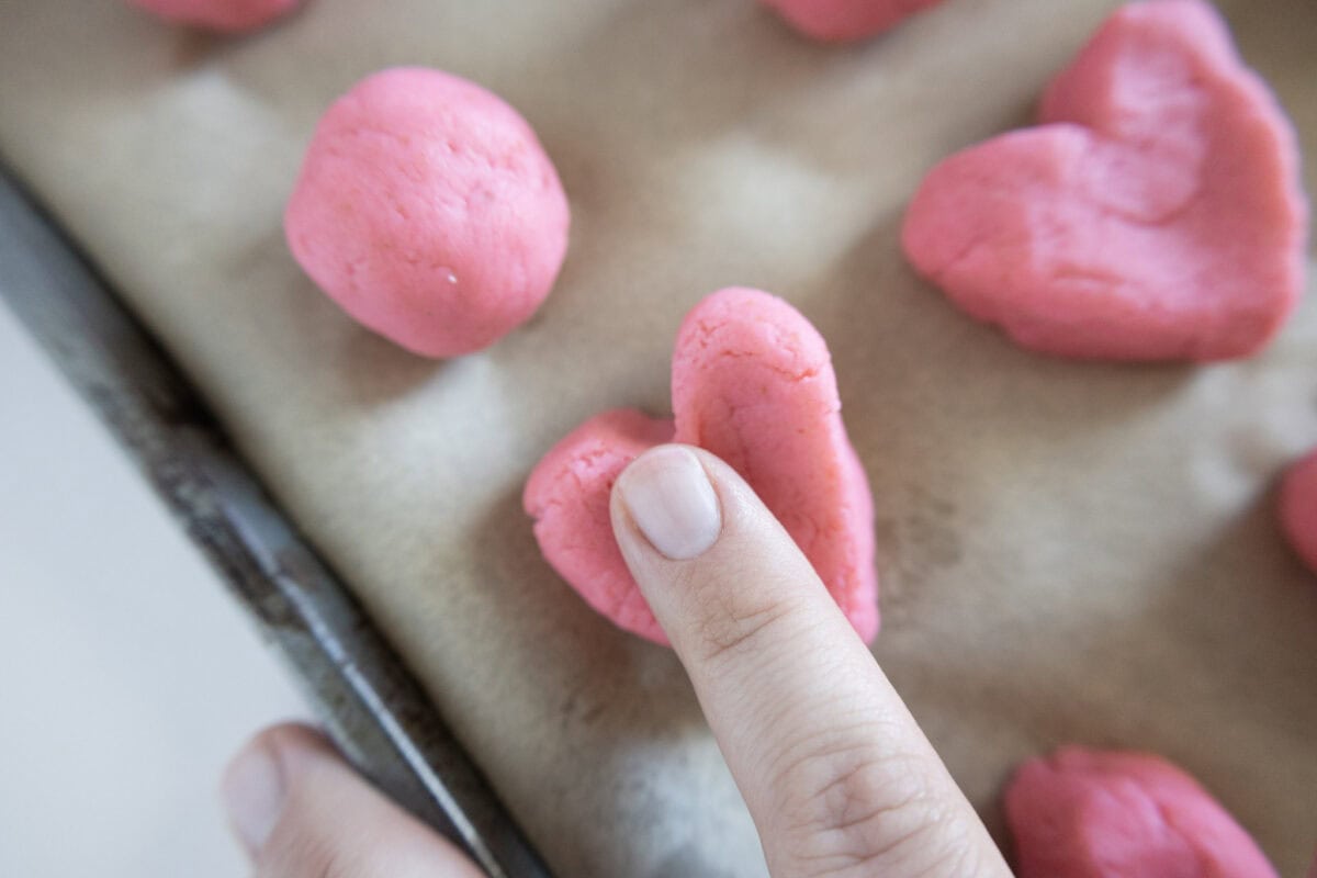 A hand shaping pink dough into a heart on a parchment-lined baking sheet.