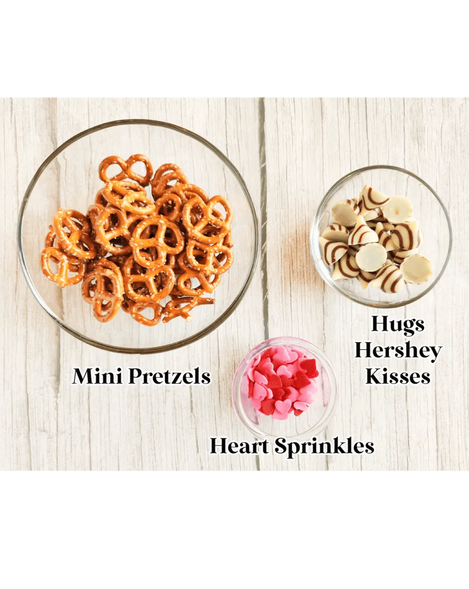 Three bowls on a wooden surface: one with mini pretzels, one with Hugs and Kisses Hershey's Kisses, and one with pink heart sprinkles.