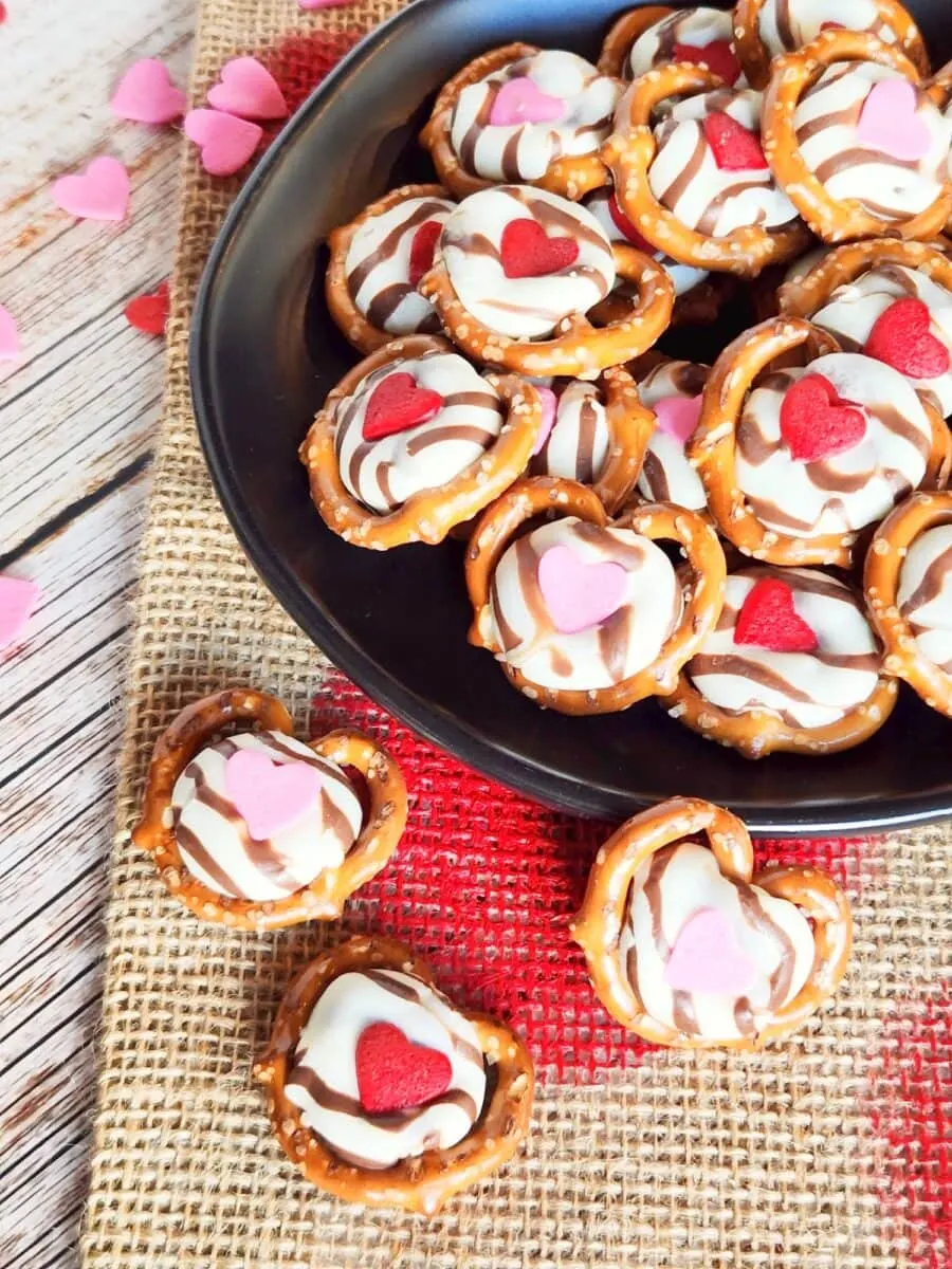 A black plate filled with chocolate-drizzled pretzels topped with pink and red heart-shaped candies, placed on a burlap mat with scattered heart confetti.