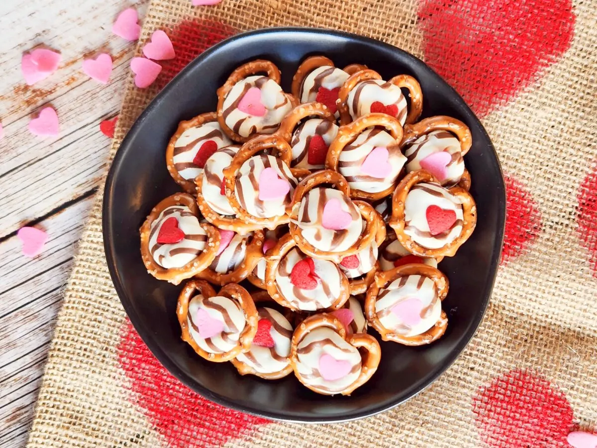 A black plate filled with heart-themed pretzel snacks topped with red and pink heart-shaped candies on a burlap cloth decorated with red hearts.