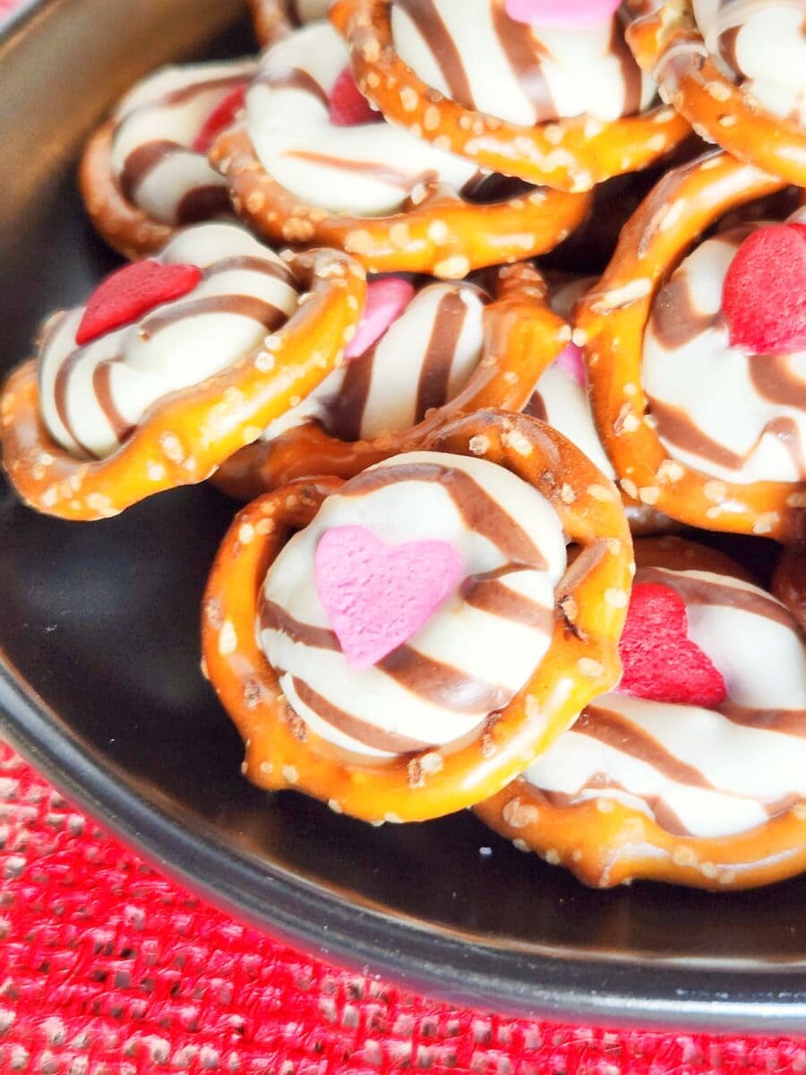 Pretzels topped with chocolate stripes, white candy, and pink heart decorations on a black plate.