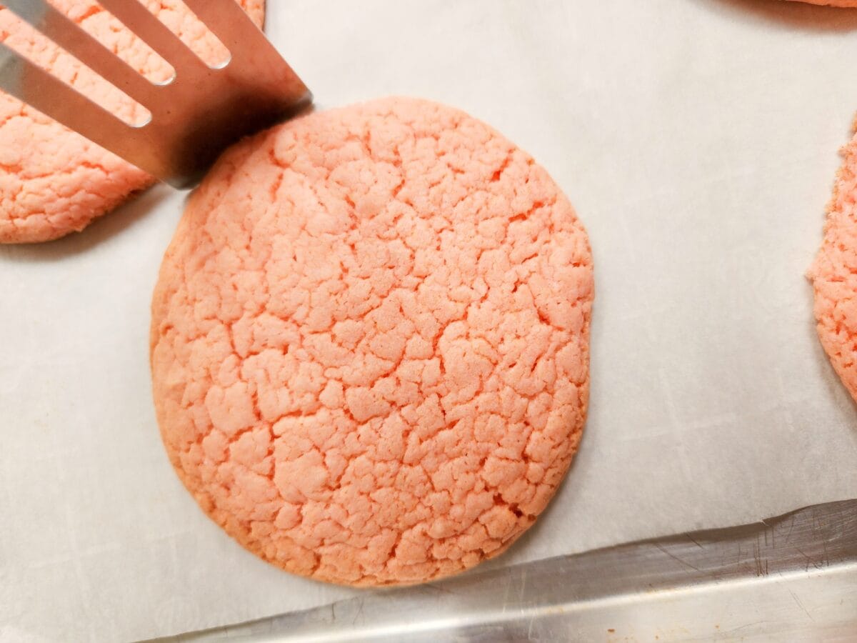 A spatula lifts a pink cookie on a baking sheet.