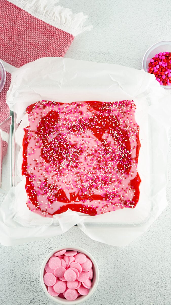 A baking tray with a pink and red dessert covered in colorful sprinkles on parchment paper. Surrounding the tray are bowls of pink candy and sprinkles on a light-colored surface.