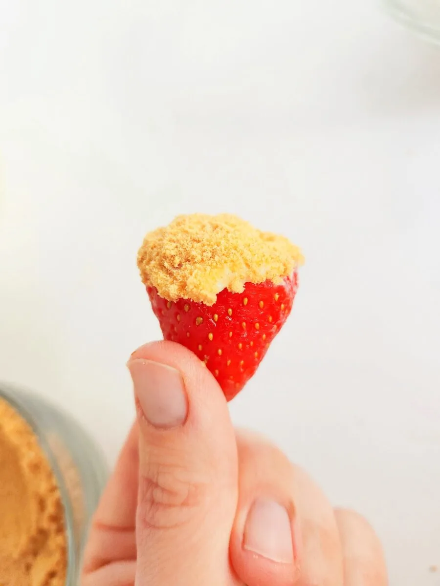 Hand holding a strawberry dipped in crushed cookies on a white background.