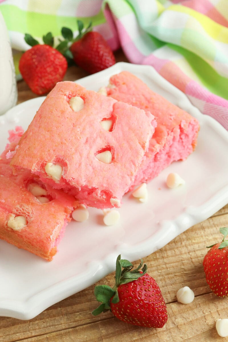 Pink strawberry cake squares with white chocolate chips on a white plate, surrounded by fresh strawberries and a colorful striped cloth.