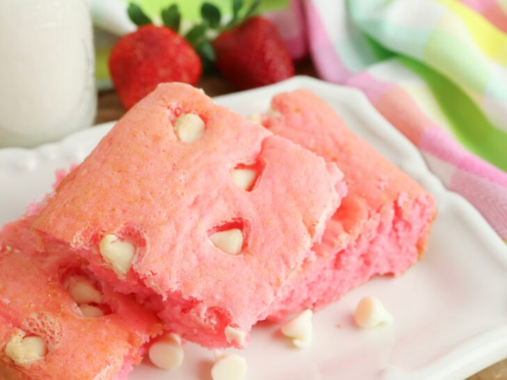 Three pink cake squares with white chocolate chips on a white plate, surrounded by strawberries and a colorful striped cloth.