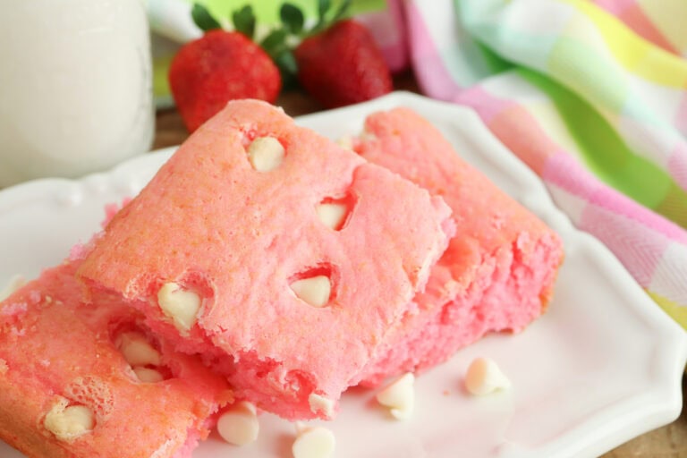 Three pink cake squares with white chocolate chips on a white plate, surrounded by strawberries and a colorful striped cloth.
