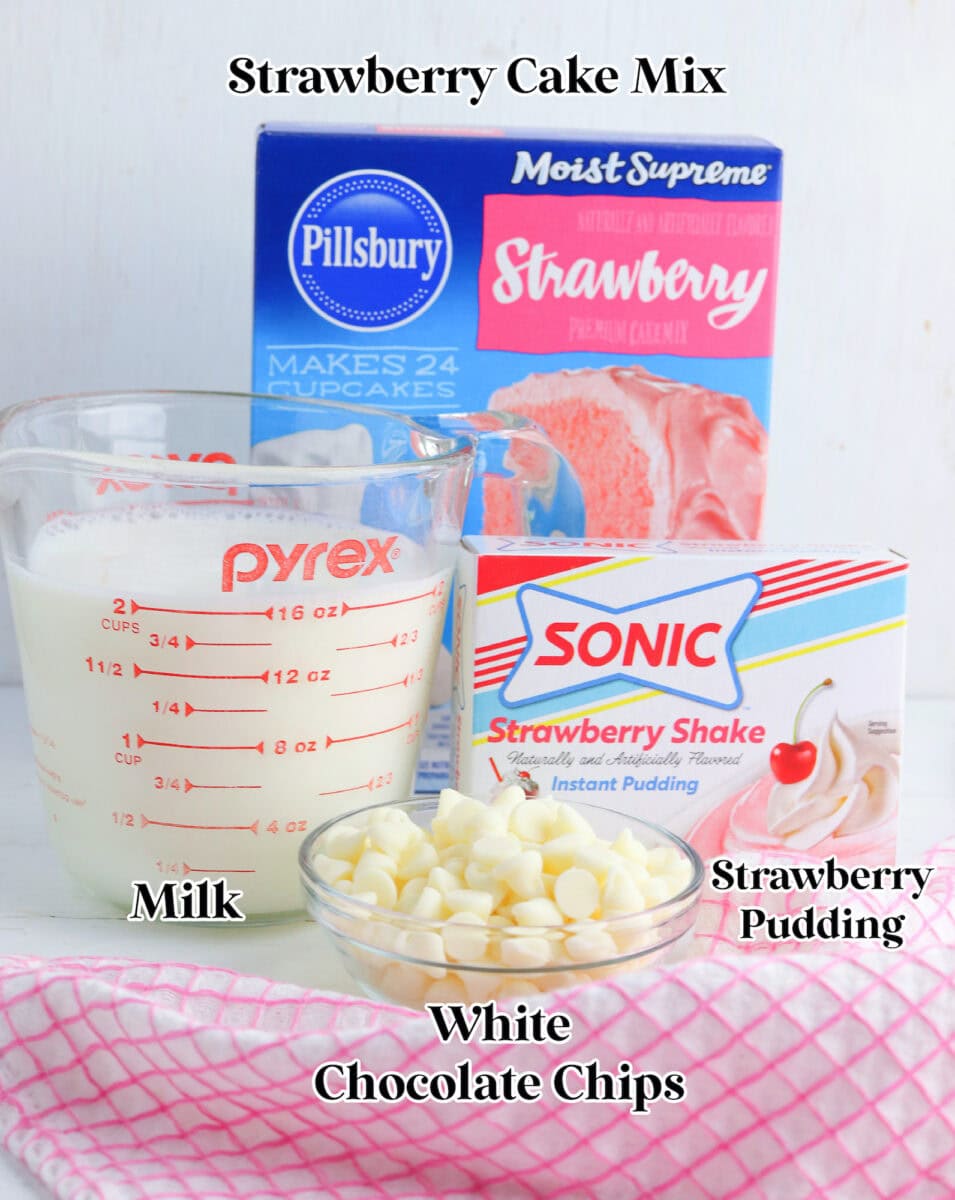 A box of strawberry cake mix, a measuring cup with milk, a box of strawberry pudding mix, and white chocolate chips arranged on the table, ready to create delectable strawberry brownies.