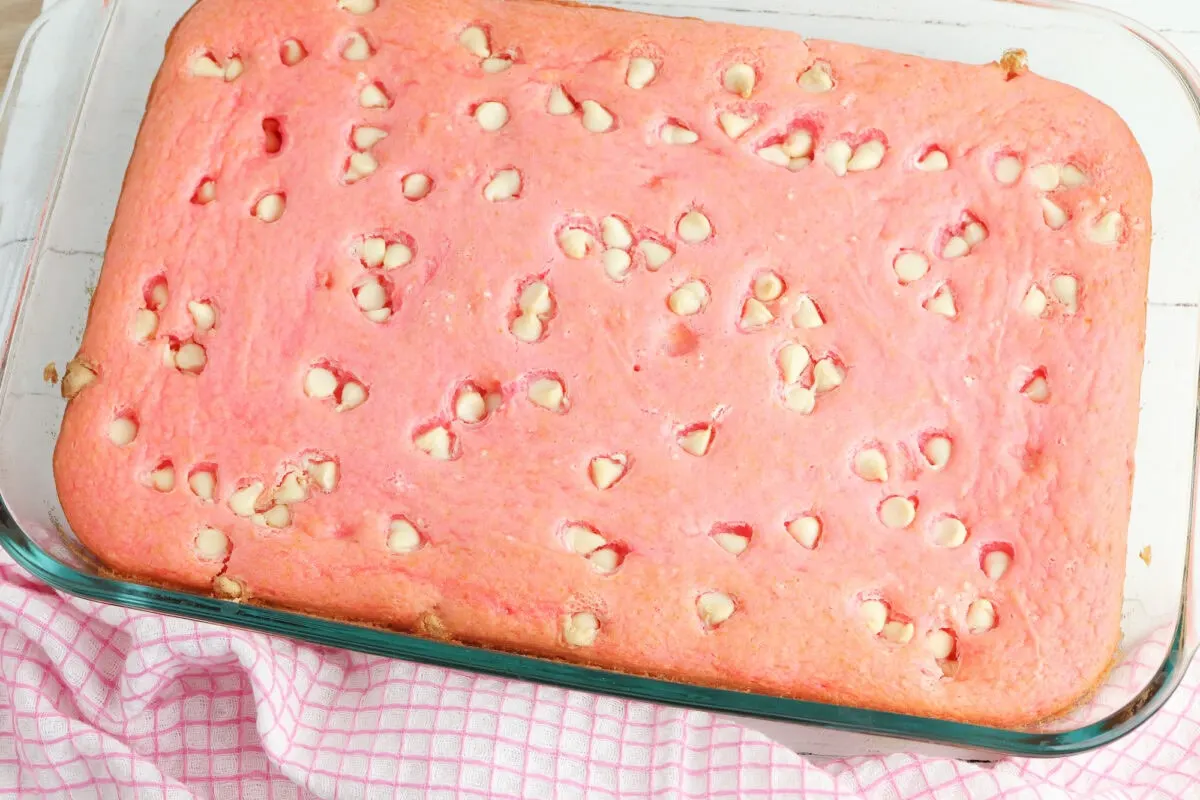 A rectangular pink cake with white chocolate chips in a glass baking dish, resting on a pink and white checkered cloth.