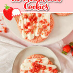 Two strawberry shortcake cookies with cream drizzle and crunchy toppings on plates, set on a cloth with fresh strawberries nearby.