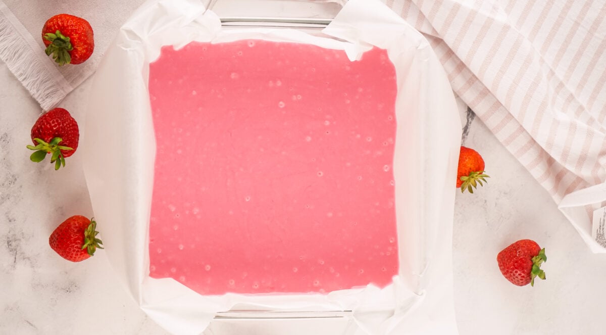 A baking dish lined with parchment paper and filled with pink batter, surrounded by strawberries on a marble countertop.